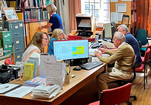 members sitting discussing around a table in an office full of cabinets, files, and book filled shelves.
